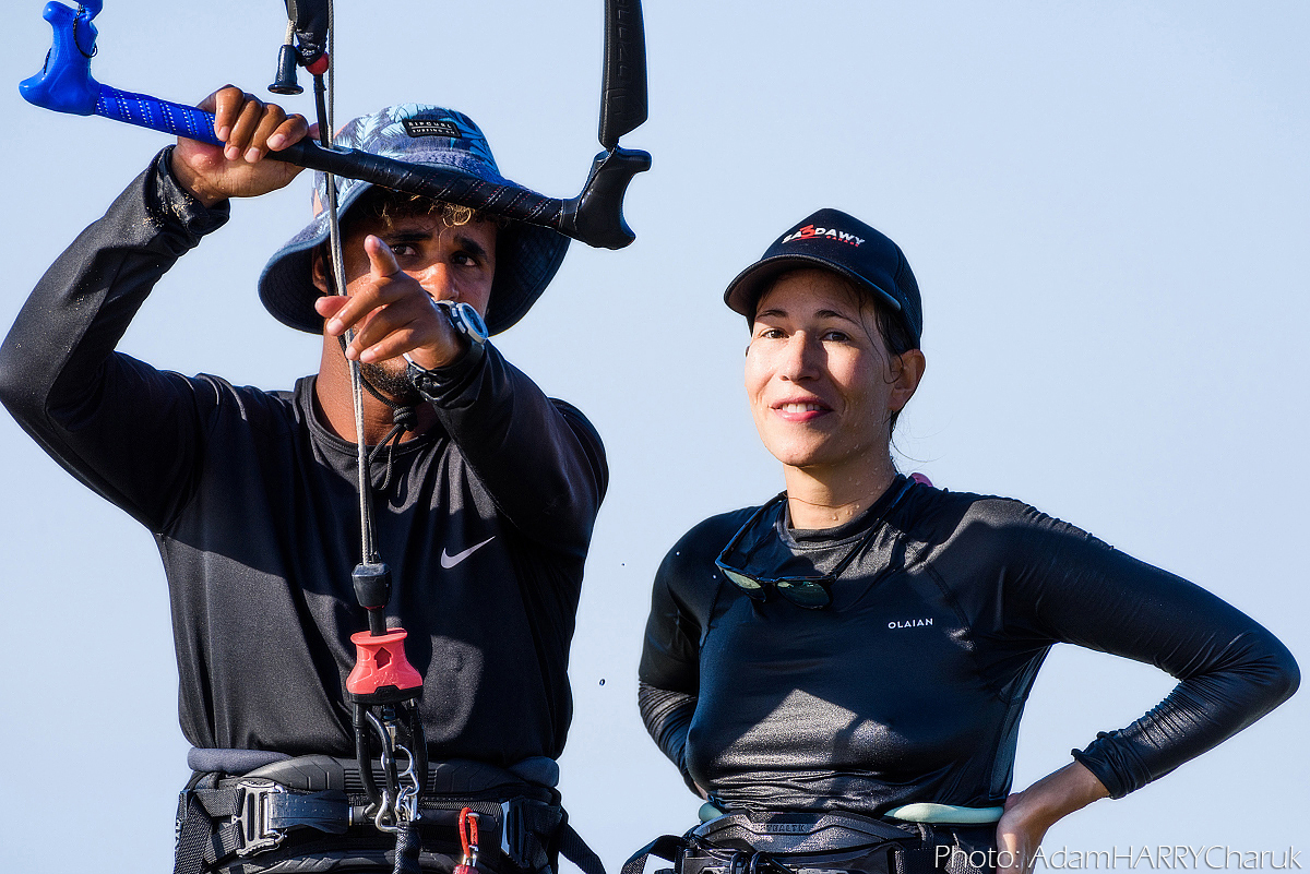 A kitesurfing instructor points out something downwind to his student. The instructor is casually holding the kite’s control bar, flying the kite at 12 o’clock above his head