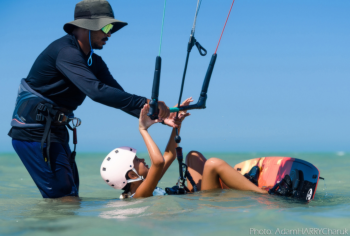 A kitesurfing student floats on their back, feet in their board’s footstraps and hands on the bar. Their teacher is behind them, giving them instructions in preparation for a waterstart