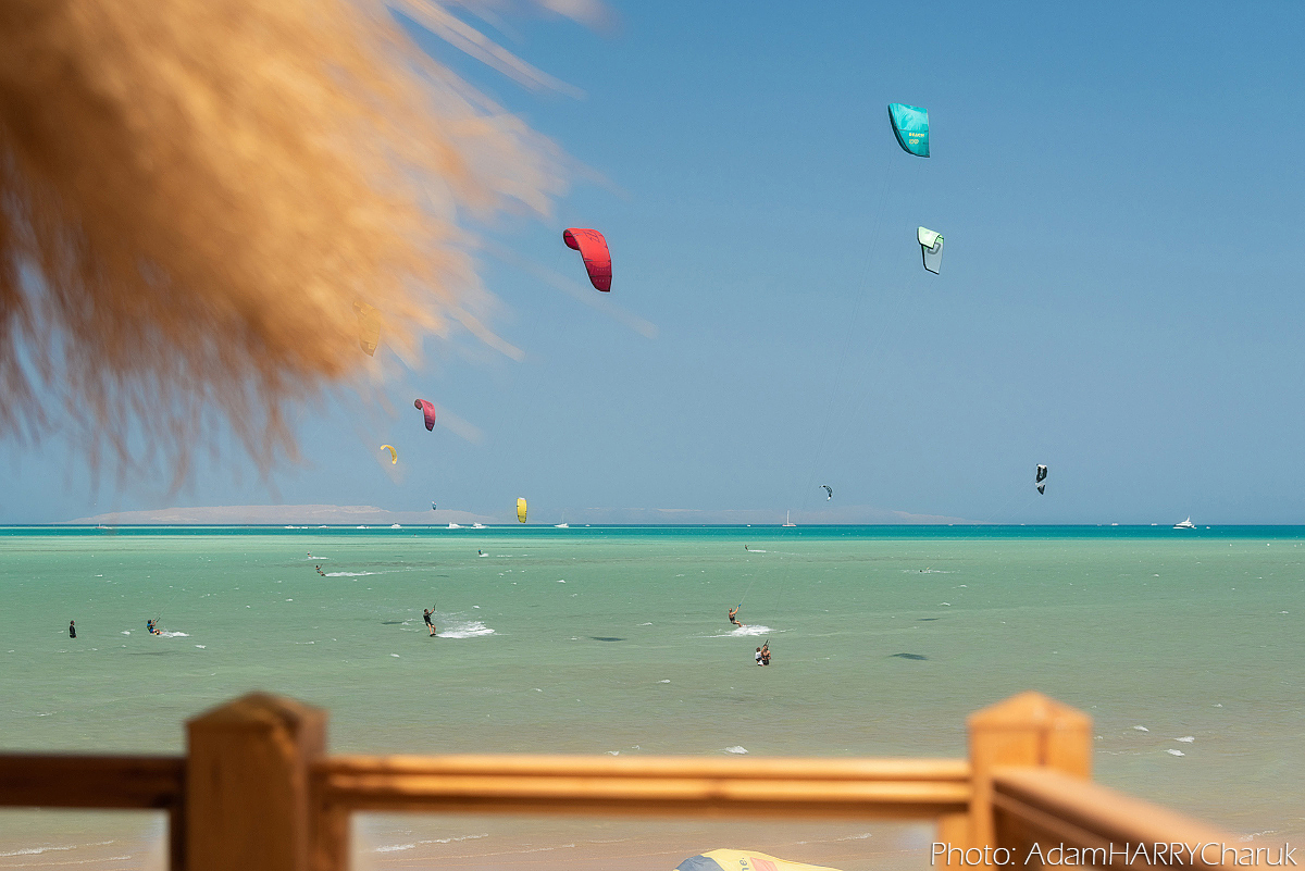 Kitesurfers in a turquoise blue bay. Looking out from a deck or viewing area, there are kitesurfers both riding, bobbing in the water and learning in the shallows. Kites of all colours are in the sky. Three in particular in the top right of the image
