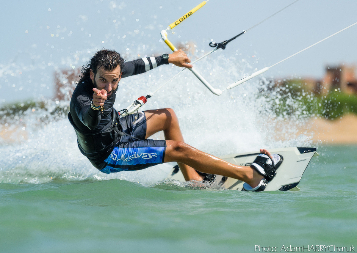 A kitesurfer is riding past the photographer, but is squatting low and pointing their index finger straight to the camera. They appear to be riding fast